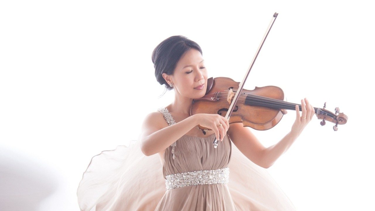  Violist Hsin Yun Huang, an Asian woman with dark brown hair styled in an updo, wears a flowing beige gown and plays her instrument in front of a white background.