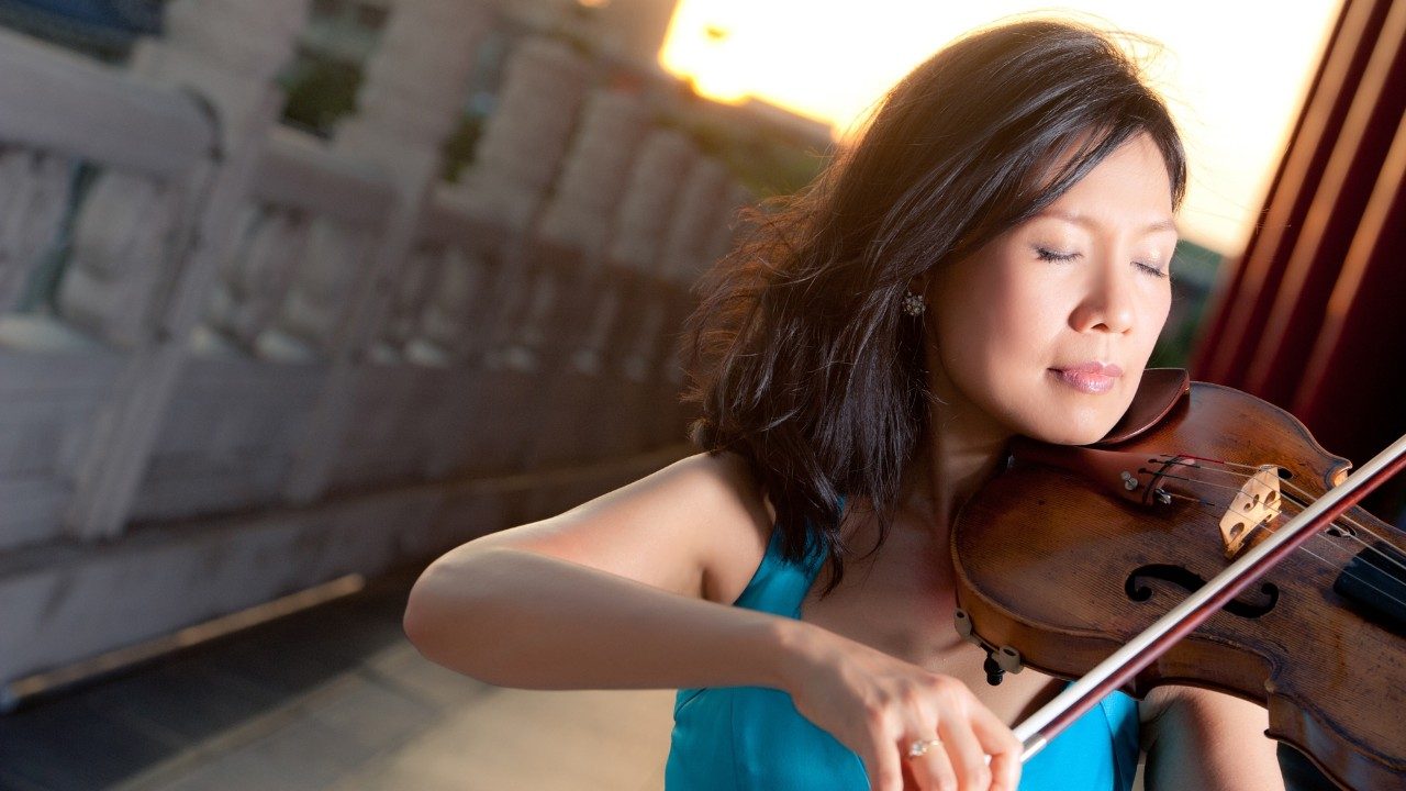  Violist Hsin Yun Huang, an Asian woman with medium length dark brown hair, plays her instrument on a rooftop, the setting sun behind her. She closes her eyes with the music and wears an electric blue tank top.