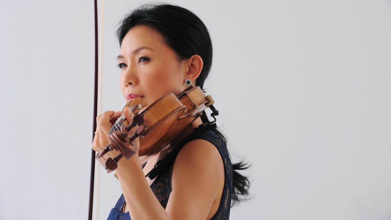 Violist Hsin Yun Huang, an Asian woman with medium length dark brown hair pulled into a ponytail, lifts her instrument to her chin in front of a pale grey background. She wears a dark blue tank top.