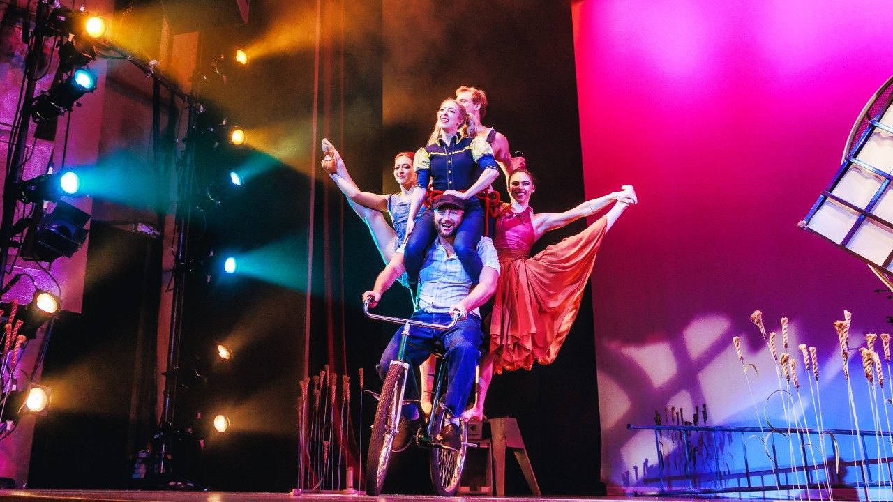  Five cast members of Cirque Mechanics ride on a green bicycle on stage, a pink backdrop visible behind them.