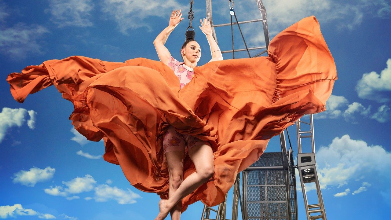 A white woman hangs by her dark brown hair from a machine of some sort, her flowing orange skirt filling the frame in front of a blue sky peppered with puffy white clouds.