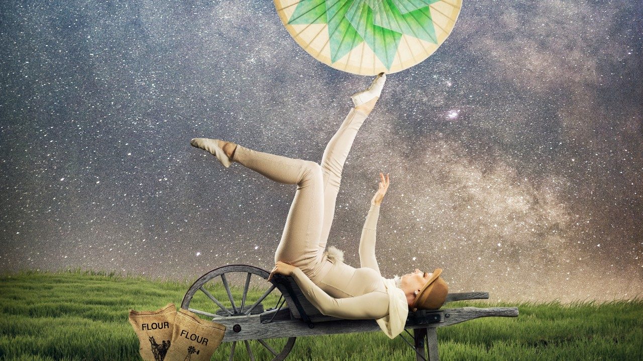  A woman in a white leotard and brown hat lays back on a wooden wheelbarrow base and twirls a white and green parasol on her feet. She is in a green field under a starry night sky.