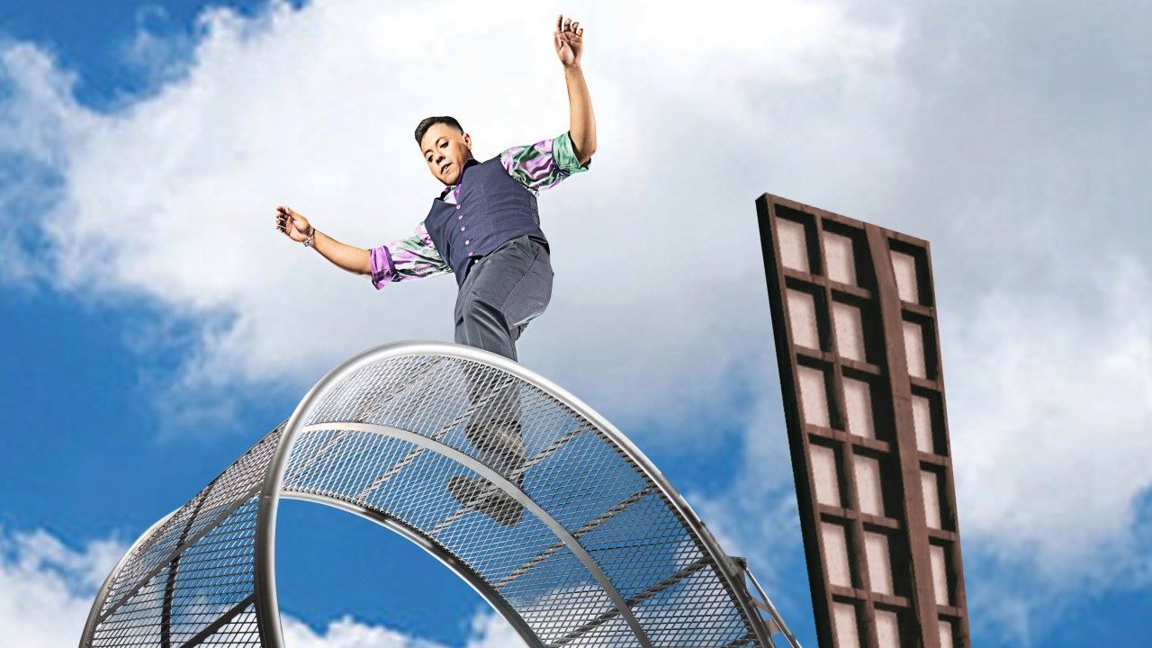  A brown man in a navy vest and purple and green button down shirt is photographed from below as he performs on a large windmill-like object with rotating circular parts. Above and behind him is a blue sky with puffy white clouds.
