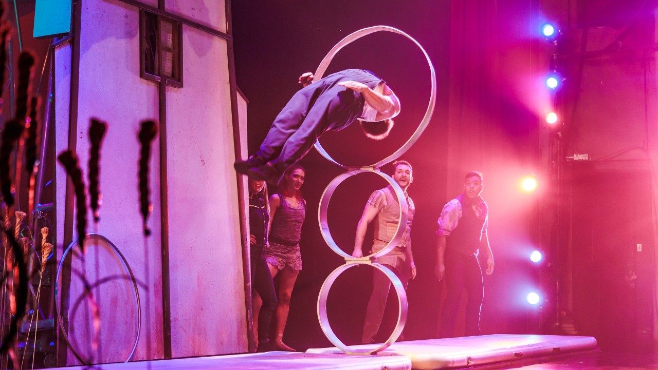  A man leaps backwards through the highest of three rings on stage, awash in pink light as his fellow cast members look on in awe.