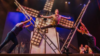  A man does the splits between two wooden ladders, which are being propped up by two fellow cast members. A large windmill is visible in the background.