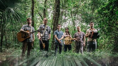  Members of the Cajun band Lost Bayou Ramblers, a group of six young white men wearing casual clothes, hold their instruments and are surrounded by the greenery of the bayou.