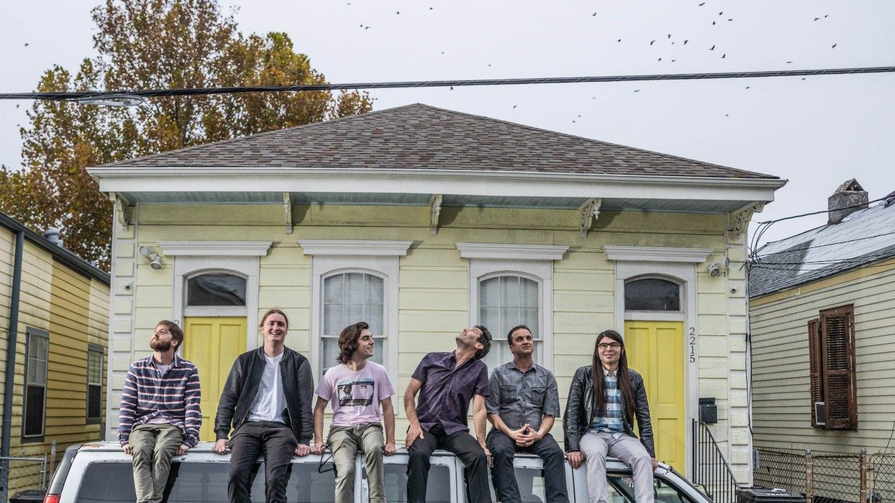  Members of the Lost Bayou Ramblers, six young white men, sit on top of a white 11-passenger van outside of a yellow shotgun-style house in New Orleans. A flock of birds passes overhead, and two of the members look up at them as they pass.