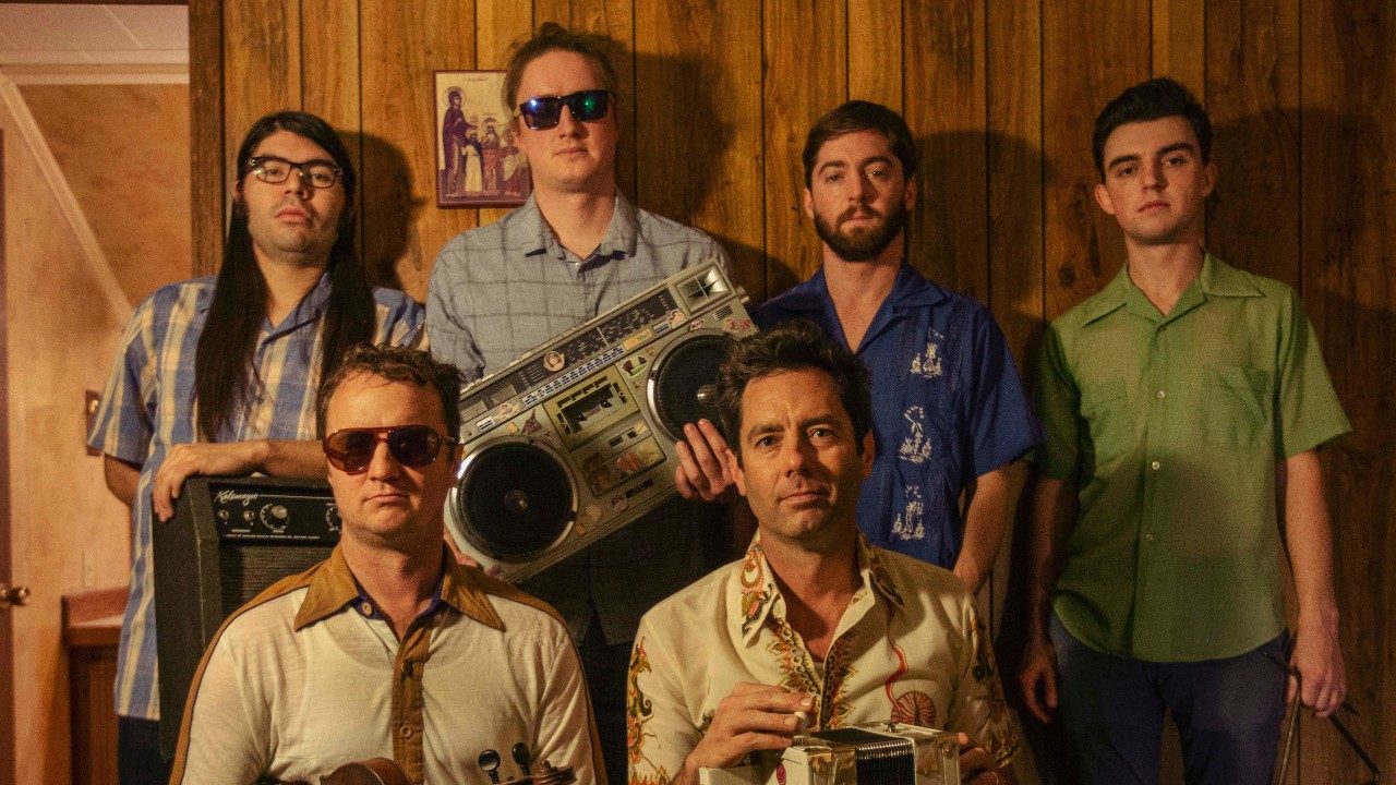  Members of the Lost Bayou Ramblers, six young white men, stand or sit in the living room of a house with wood paneling. They wear casual, '70s-looking clothes, and hold instruments, amps, and boom boxes.