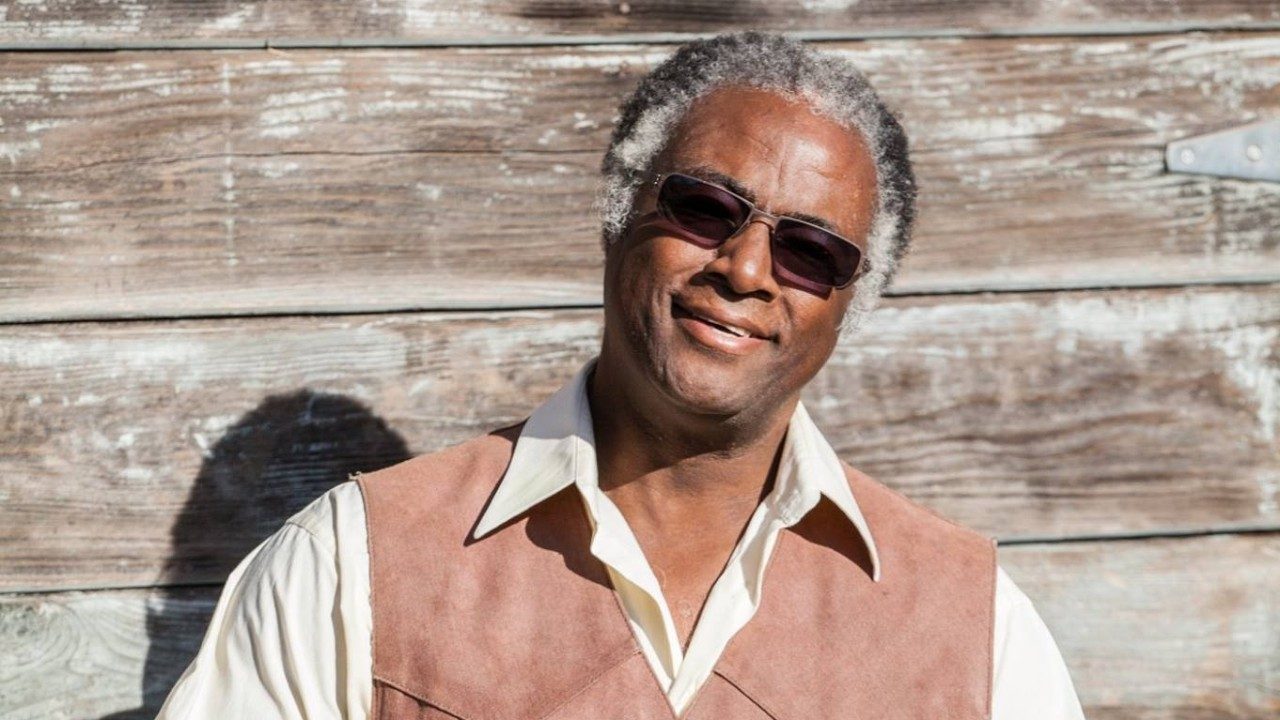 Old-time musician Earl White, an older Black man with salt and pepper, medium length natural hair, wearing a light brown suede vest over a white button down shirt. He stands in front of a weathered wood plank exterior wall.