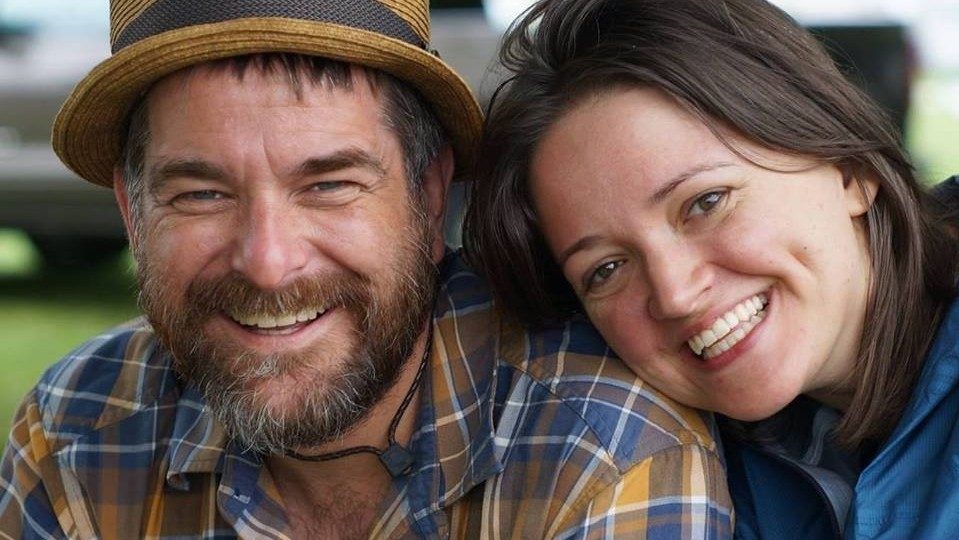 The members of the Lovely Mountaineers, a white man in a blue and yellow plaid shirt and straw hat, and a woman in a blue jacket with dark brown hair. She leans her head on his shoulder as they both smile towards the camera.