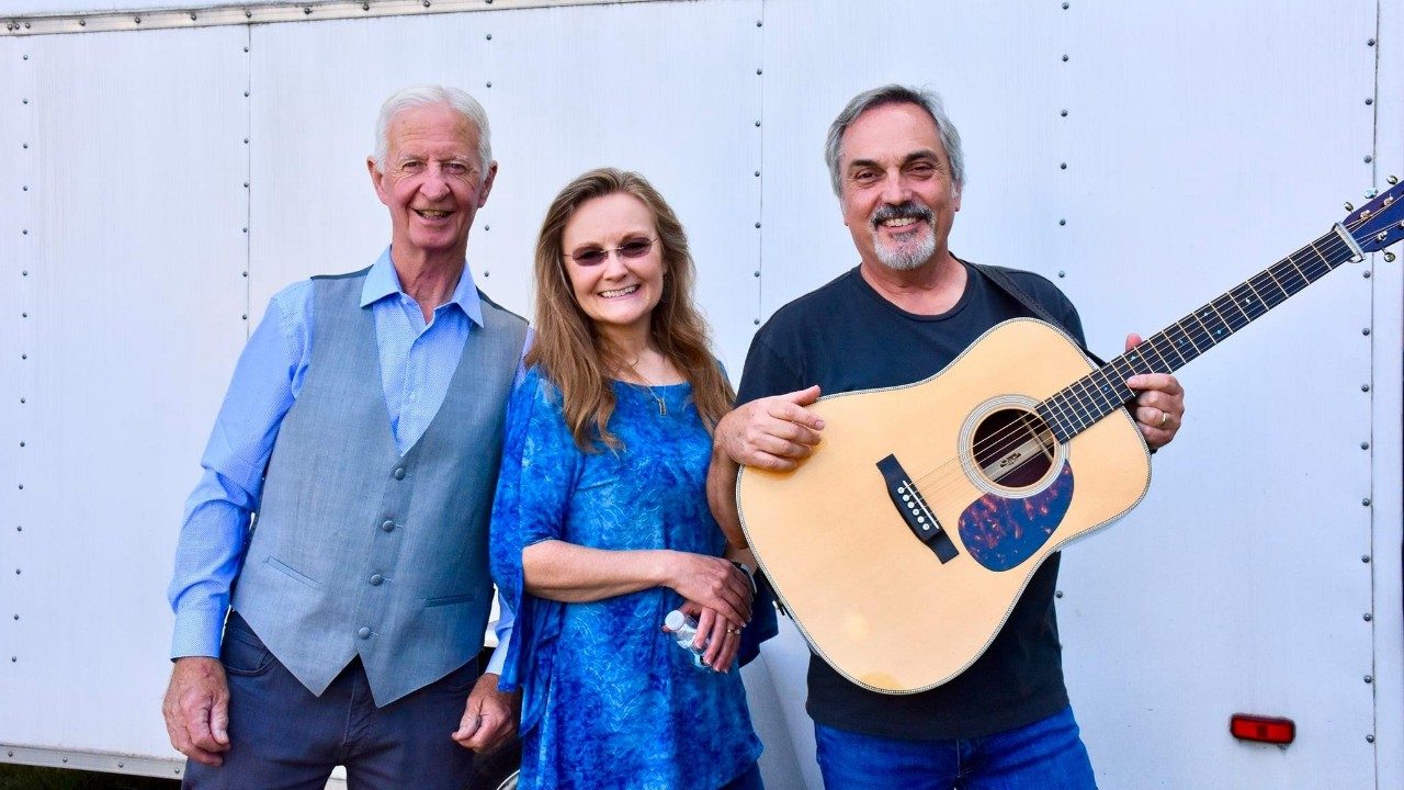 The members of Shelton and Williams, two older white men and one white woman, stand in front of a white truck trailer, one man holding an acoustic guitar.