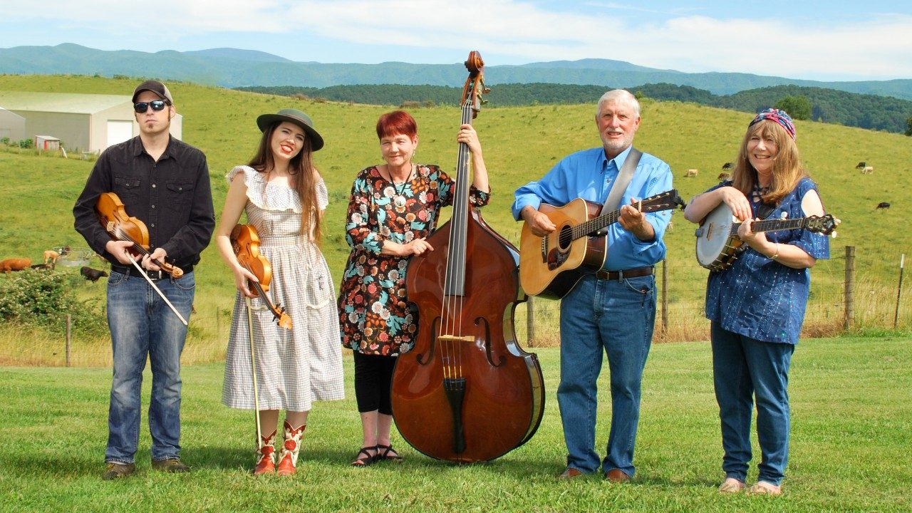  The members of the Whitetop Mountain Band hold their instruments and stand in a green field, mountain ranges visible in the distance behind them. From left, a young white man holding a fiddle in a black button down shirt and jeans, a young white woman with long brown hair holding a fiddle and wearing a light pink dress, a middle aged white woman with short red-brown hair and holding a double bass, an older white man with white hair holding an acoustic guitar and wearing a blue button down shirt and jeans, and a middle aged white woman with medium length brown hair holding a banjo and wearing a denim shirt and denim pants.