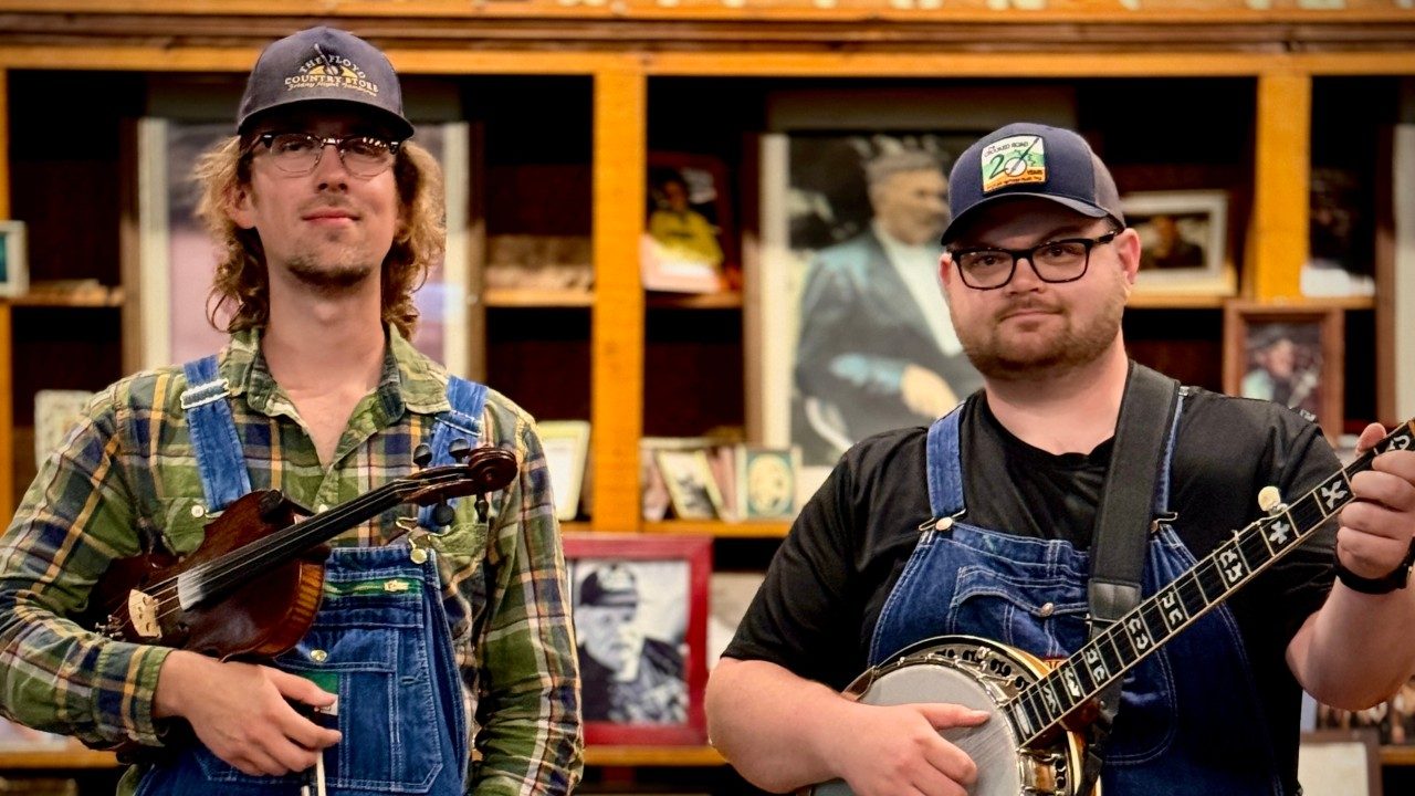 Left, Raistin Brabson, and right, Jared Boyd, two young white men wearing denim overalls, and hats, and holding a fiddle and banjo, respectively.