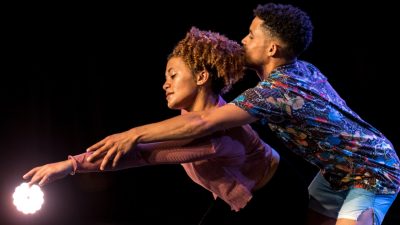  Two dancers perform "Krummelpap, Scandals Wrapped with Prayer" on stage. A Black woman, at left, and a Black man, at right, extend their left arms out towards a stage light. She has honey blonde natural curly hair pulled into a ponytaile and wears a pink long sleeved shirt. He has medium length natural hair and wears a brightly colored patterned shirt and light blue shorts.