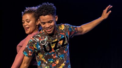  A young Black woman, left, and a young Black man, right, dance together on stage during "Krummelpap, Scandals Wrapped with Prayer." She has honey blond natural hair pulled into a ponytail, wears a pink shirt, and sticks her tongue out through a flirty smile. He wears a brightly colored patterned shirt, on which black letters read "UZZI," and extends his right arm out gracefully.
