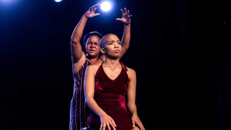  Two light skinned Black women, one middle aged, in the background, and one younger, in the foreground, perform "Krummelpap, Scandals Wrapped with Prayer" on stage. The younger woman has a shaved head and wears a wine-colored velvet halter neck dress and sits on a stool. The older woman wears a striped tank top dress and stands behind her, her face twisted in concentration and both hands raised above the head of the younger woman, as if in prayer. Between her hands, a bright stage light is visible.