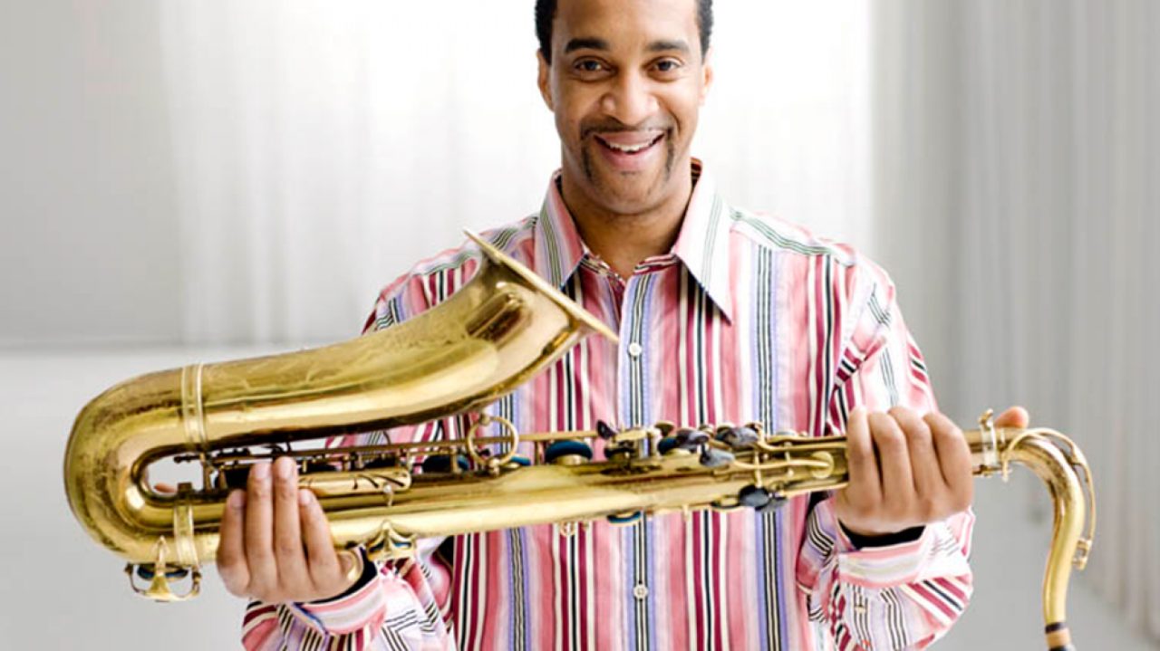  Jazz saxophonist Javon Jackson, a Black man with short natural hair, holds his saxophone sideways in both hands in front of his body. He wears a pink and blue striped button down shirt and smiles towards the camera.