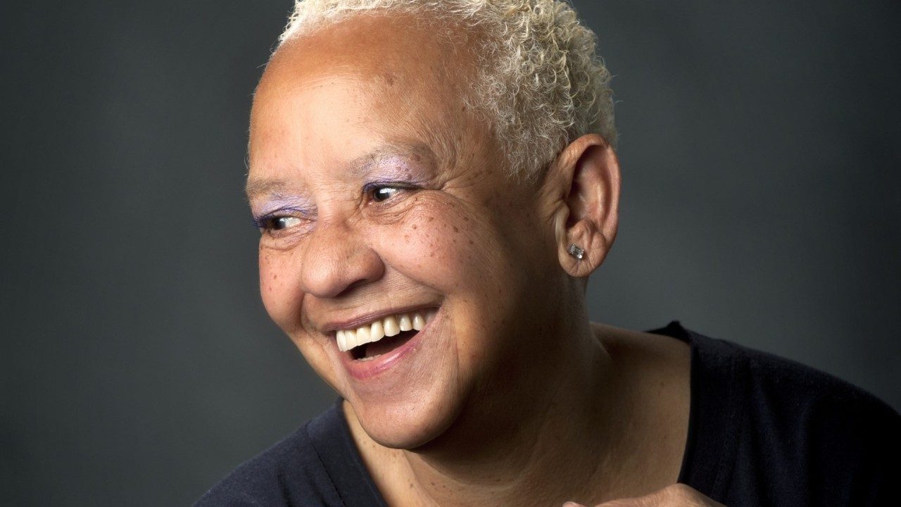  A headshot of Nikki Giovanni, an older Black woman with short, bleached blonde, curly hair. She wears a black shirt and purple eyeshadow and smiles widely, looking away from the camera in front of a grey background.
