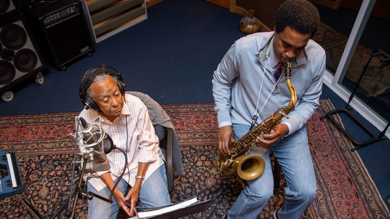 At left, Nikki Giovanni, an older Black woman with short, grey, natural hair, and at right, Javon Jackson, a Black man with a short Afro playing a saxophone. They sit in chairs in a recording studio. She wears headphones and reads from a music book while he plays.
