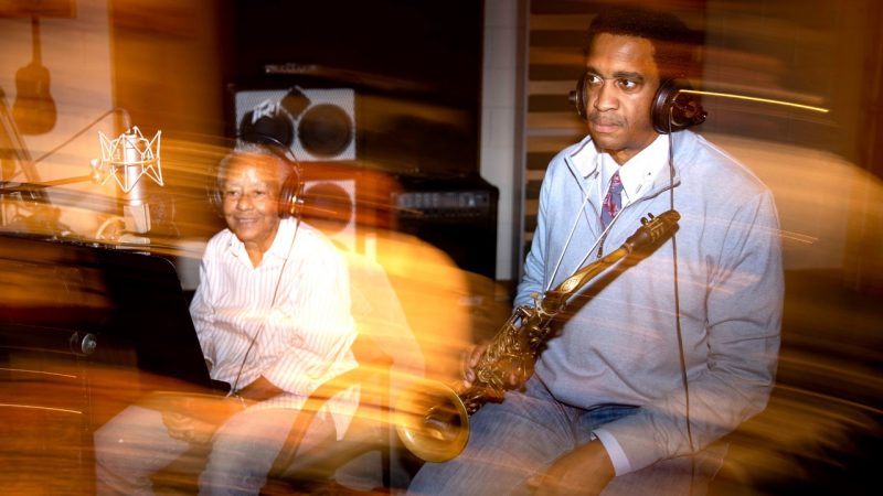  At left, Nikki Giovanni, an older Black woman with short, grey, natural hair, and at right, Javon Jackson, a Black man with a short Afro. They sit in a recording studio, both wearing headphones, and he holds his saxophone. Blurred yellow lights reflect in front of the pair.