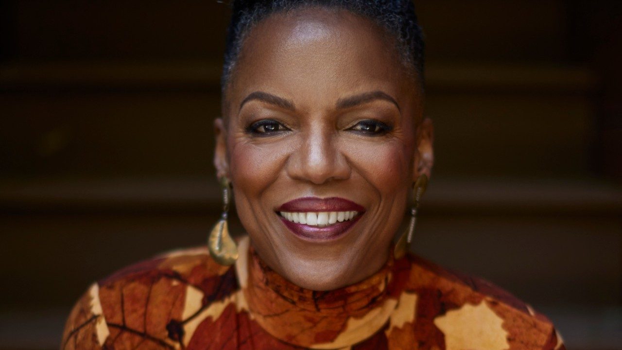  Jazz singer Nnenna Freelon, a middle aged Black woman wearing a turtleneck dress with a print that looks like red, orange, and yellow fall leaves. She smiles towards the camera wearing a wine-colored lipstick.