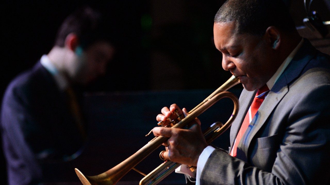  Trumpet player Wynton Marsalis, a Black man with short natural hair, plays his trumpet in the foreground, a pianist is visible but out of focus in the background. Marsalis wears a dark grey suit, white button down shirt, and red and white striped tie.