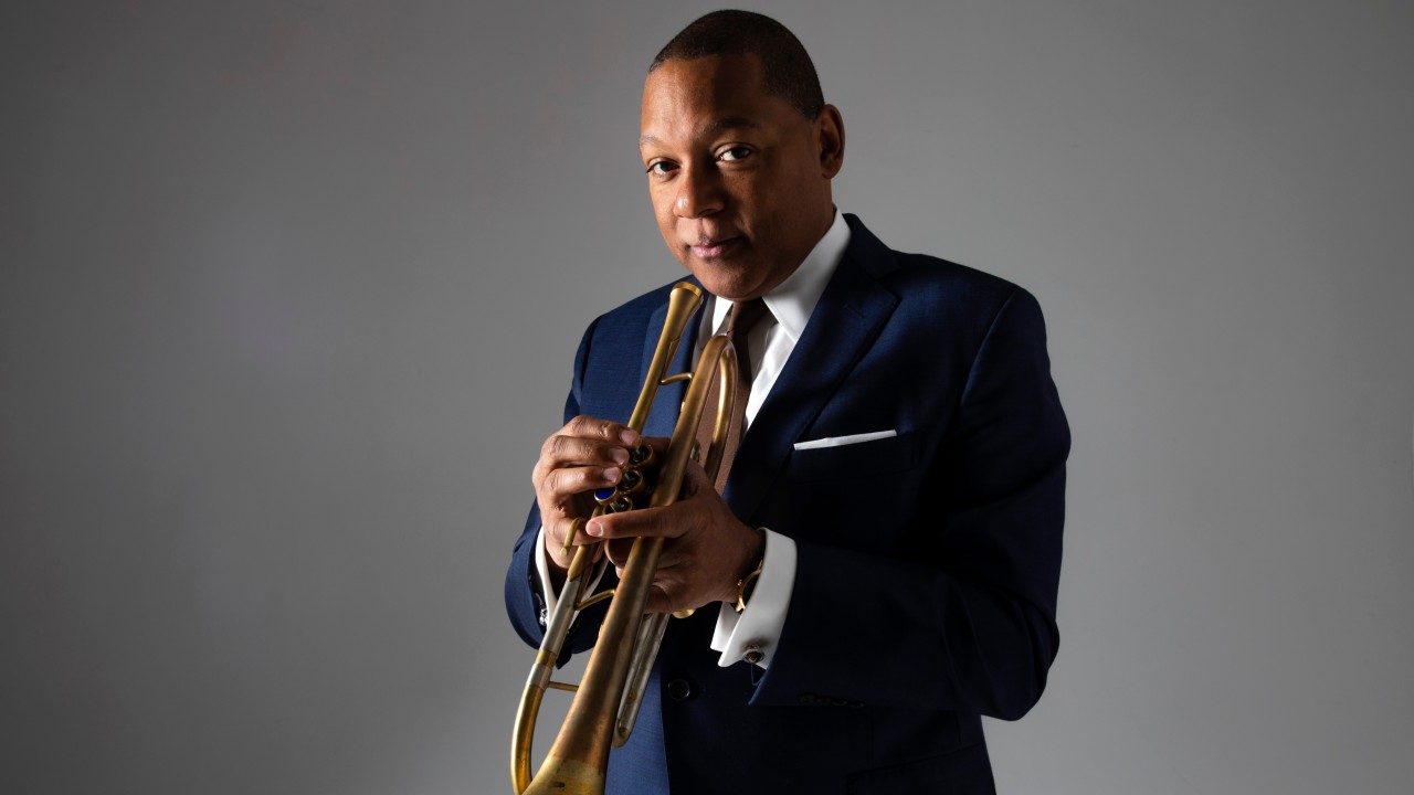  Trumpet player Wynton Marsalis, a Black man with short natural hair, wears a navy blue suit and holds his instrument in this headshot, taken against a grey background.