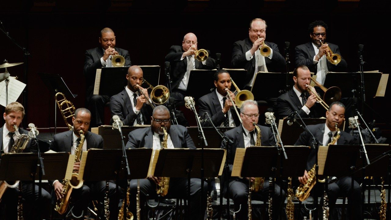  Members of Jazz at Lincoln Center Orchestra perform on stage wearing black suits. Pictured are 12 men, five white and seven Black.
