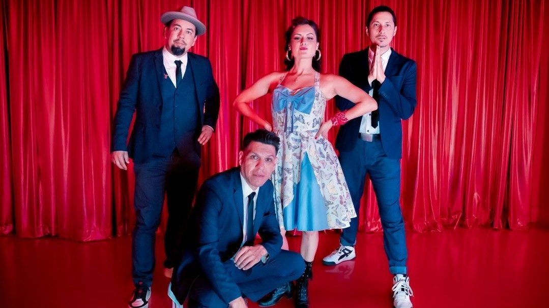  Four members of East Los Angeles Chicano indie folk band Las Cafeteras pose in front of a red curtain. The three men wear navy blue suits and ties, while the one woman, in the middle, wears a rockabilly-inspired A-line floral halter dress.