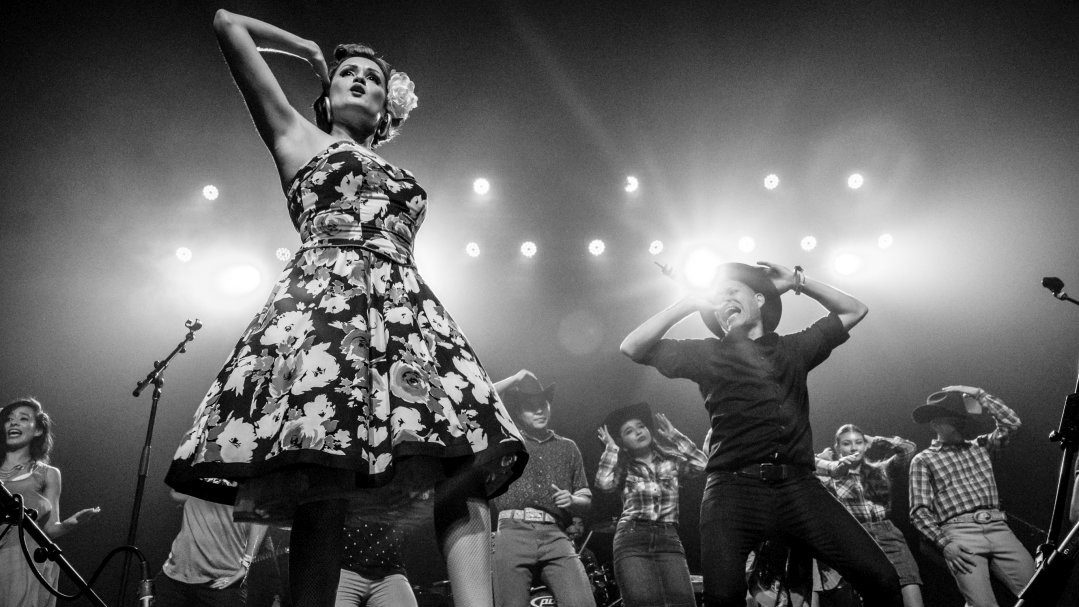  East Los Angeles Chicano indie-folk band Las Cafeteras performs on stage in this black and white photo. In the foreground, the female lead singer wears a roackbilly-inspired floral A-line halter dress, large thick gold hoops, and a large white flower tucked behind her ear. Behind her, men and women in western attire dance and sing.