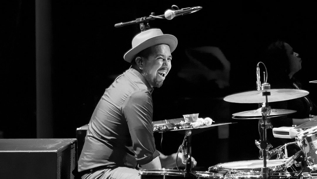  The drummer for East Los Angeles Chicano indie-folk band Las Cafeteras, a brown man in a button down shirt and hat, sits at his drum set and smiles widely in this black and white image.