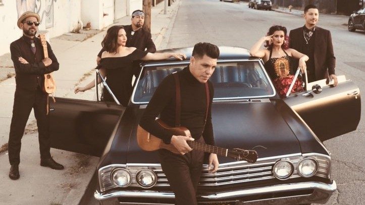  Members of East Los Angeles Chicano indie-folk band Las Cafeteras pose around a restored matte black El Camino. In the center foreground, the lead singer holds a Mexican ukulele-sized guitar. 
