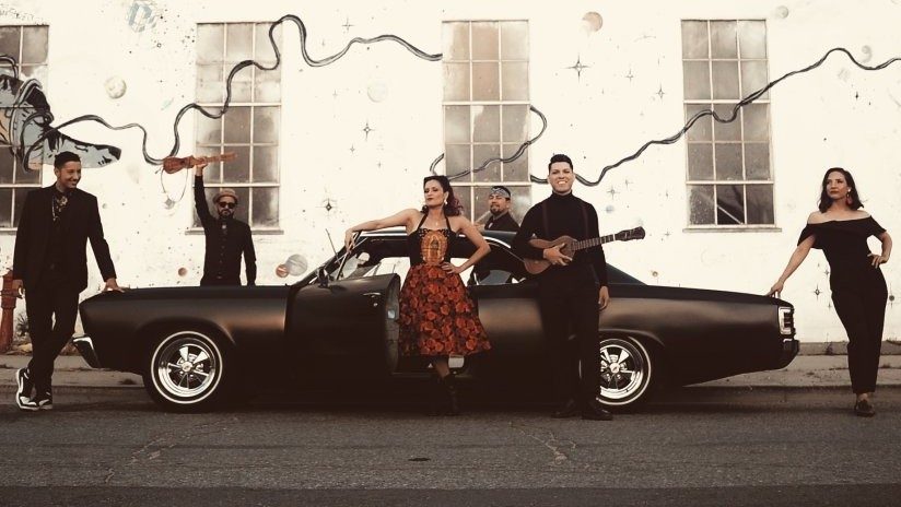  Members of East Los Angeles Chicano indie-folk band Las Cafeteras pose around a restored matte black El Camino. Some members hold Mexican ukulele-sized guitars. They pose in the street in front of a exterior wall mural.