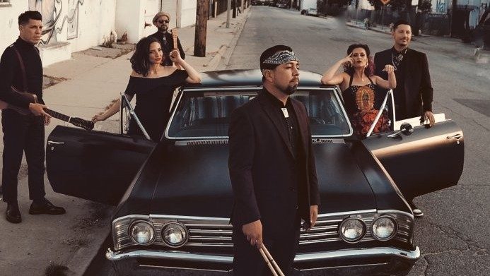  Members of East Los Angeles Chicano indie-folk band Las Cafeteras pose around a restored matte black El Camino. In the center foreground, a brown man holds a pair of drum sticks. The two brown women stand in the open car doors and the remaining three brown men stand nearby wearing all black.