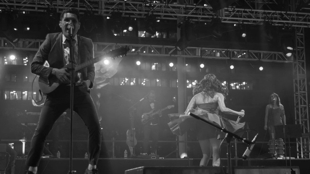  East Los Angeles Chicano indie-folk band Las Cafeteras performs on stage in this black and white photo. The members wear rockabilly-inspired clothes, the male lead singer sings into a microphone in the foreground.
