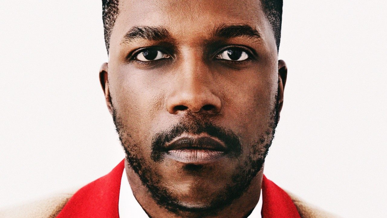  Leslie Odom, Jr., a young Black man with short natural hair and a short beard, wears a camel colored pea coat with a red collar over a white button down shirt and stares into the camera in front of a pale beige background.