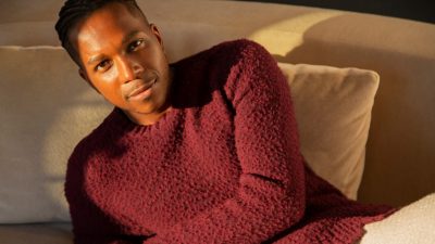  Leslie Odom, Jr., a young Black man with cornrows, wears a textured maroon sweater and white pants and reclines on a white linen couch into a ray of sunlight at golden hour. 