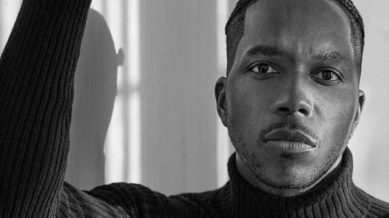  Leslie Odom, Jr., a young Black man with cornrows, wears a dark turtleneck sweater and looks into the camera. One arm is propped up and the sunlight kisses the right side of his face.
