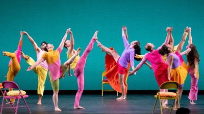  Members of Mark Morris Dance Group perform "The Look of Love" in bright, pastel, color blocked uniforms. Brightly colored metal folding chairs are peppered throughout the dancers, and the background is teal.