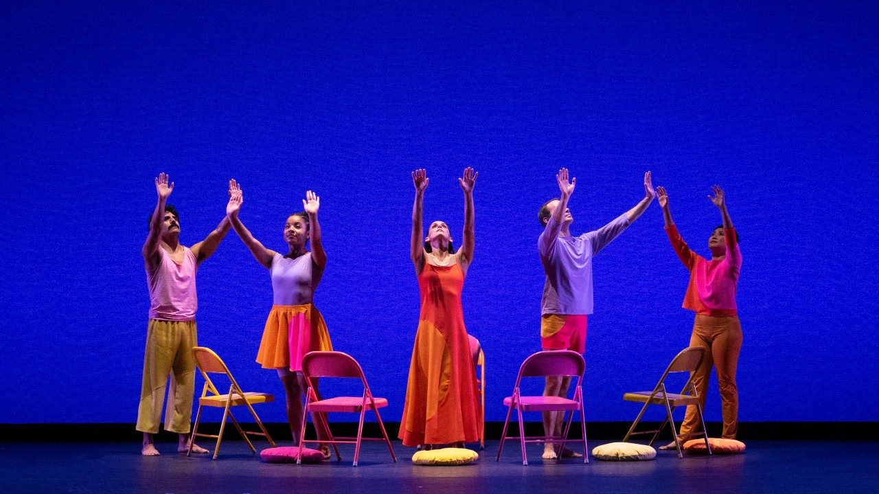  Members of Mark Morris Dance Group perform "The Look of Love," the dancers raise their hands towards the sky in front of a royal blue background. Facing them are four brightly colored metal folding chairs, and corresponding seat cushions are on the floor near each chair.