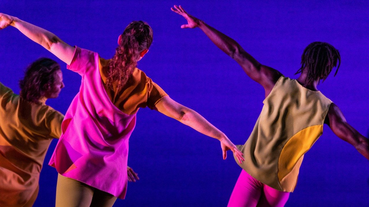  Three dancers of Mark Morris Dance Group perform "The Look of Love" on stage. They face away from the camera with arms outstretched into airplane arms, each tilted diagonally from left to right against a royal blue background.  They wear bright, colorblocked uniforms.