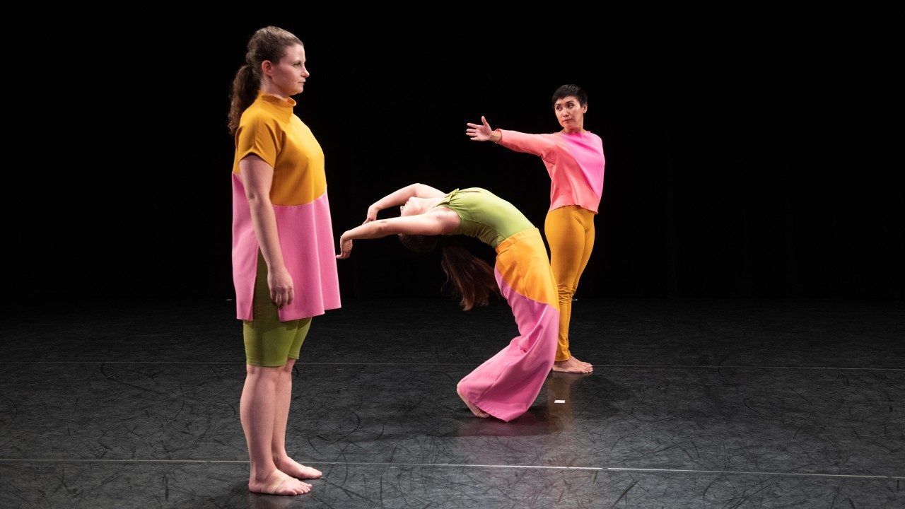  Three female dancers perform "The Look of Love" on stage against a black background, wearing bright, color blocked uniforms. At left, a white woman stands still and looks straight ahead. In the middle, a white woman bends backwards, both arms extended back behind her head. A third woman, at right, stands still and extends her arm towards the woman in the middle.