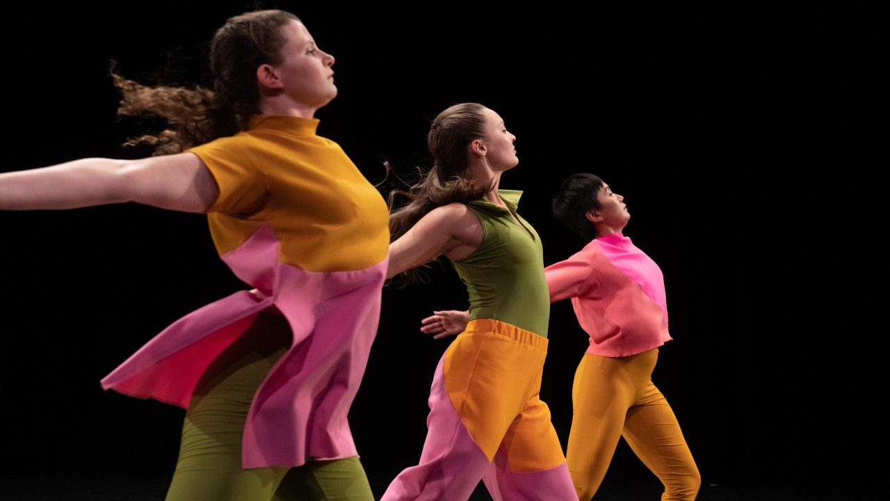  Three women dancers of Mark Morris Dance Group perform "The Look of Love" on stage, all facing towards the right edge of the frame, in front of a black background. The wear bright, color blocked uniforms.