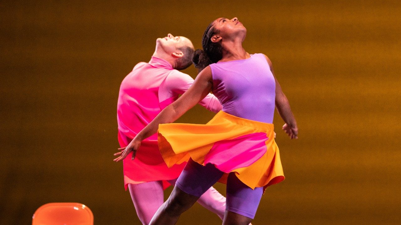  Two dancers of the Mark Morris Dance Group perform on stage in bright, colorblocked uniforms, each leaping away from the other and looking skyward.