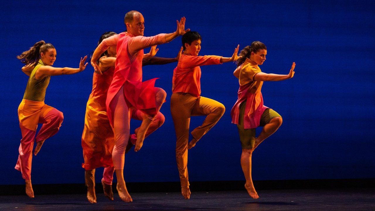  Dancers of the Mark Morris Dance Group jump with one knee bent at a right angle, one arm straight out in front of them as if to stop something from coming their way. The dancers wear bright, color blocked uniforms in front of a blue background.