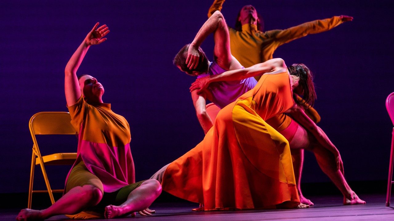  Dancers perform "The Look of Love" on stage in bright, color blocked uniforms. One woman sits on the floor, one arm raised above her head. A male and female pair leans back to back in deep lunges.