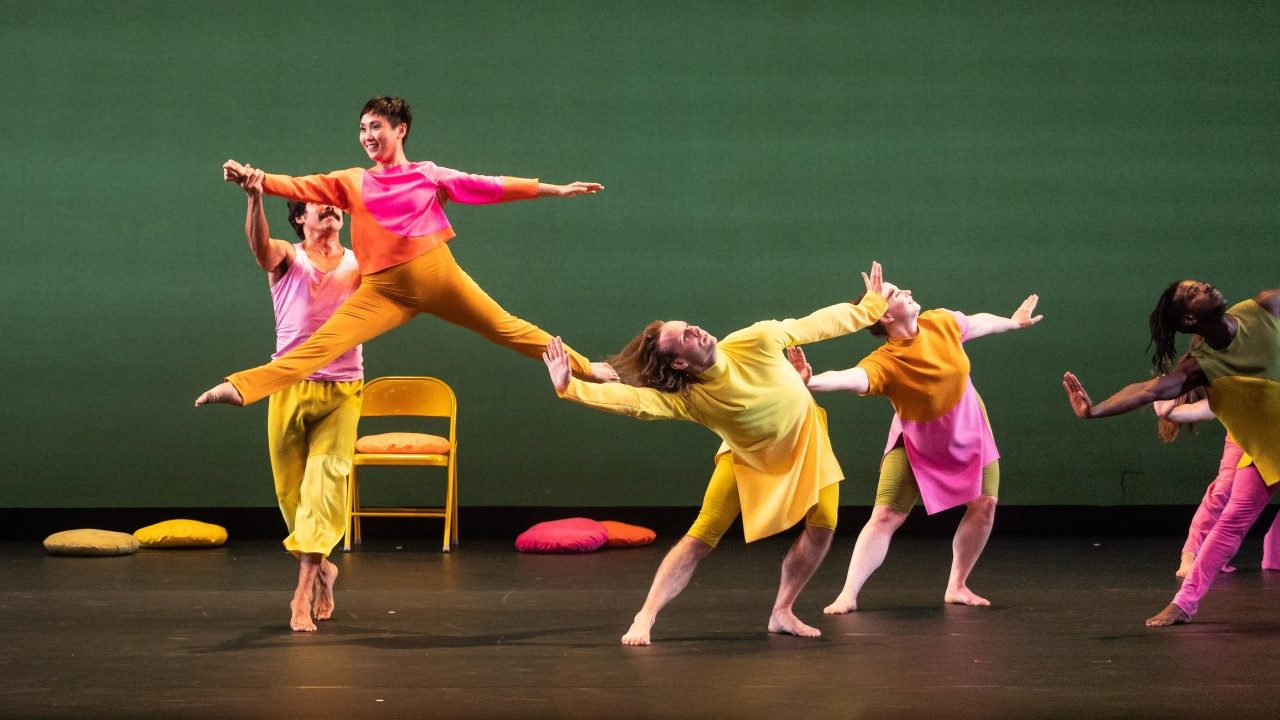  Dancers perform "The Look of Love" on stage wearing bright, color blocked uniforms. In the foreground, an Asian woman with short, dark brown hair leaps into the air, assisted by a male dancer, against a green background.