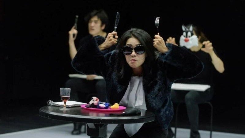  An Asian woman in a black fur coat and sunglasses sits at a round one-person table, a glass of red wine, a notebook, and a pink plate with a Mickey Mouse doll sit on the table in front of her. She lifts a fork and a knife up towards her head, almost like animal ears. Behind her, a man holds a gun and a woman wears a Sylvester the Cat mask.