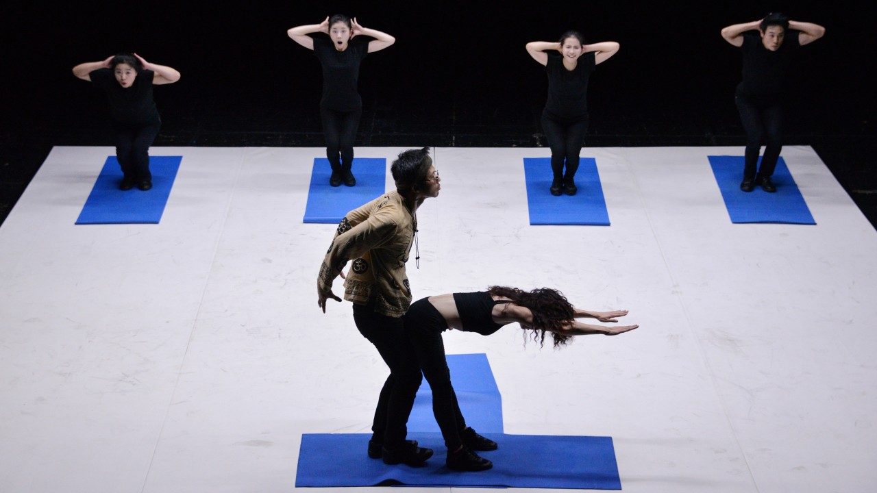  Male and female yoga instructors perform a suggestive yoga pose in a standing position on two blue yoga mats. The class, four actors dressed all in black, is in chair pose on their own blue yoga mats with their hands behind their heads and elbows open wide.
