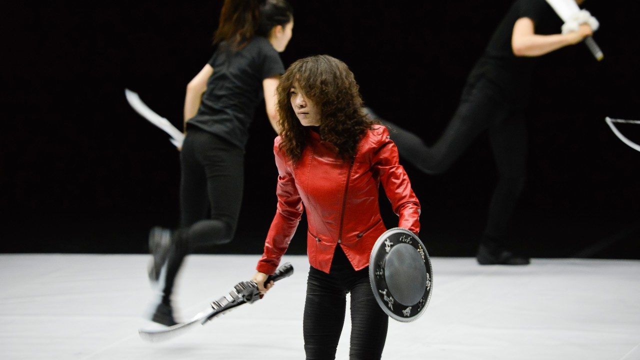 An Asian woman with long lightish brown hair wears a red leather jacket and black pants and holds a toy sword and shield. Behind her, two people with their own toy weapons and wearing all black chase each other across the stage.